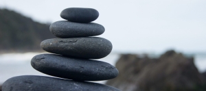 Zbliżenie na stos pięciu gładkich, ciemnych kamieni ułożonych jeden na drugim na rozmytym tle, w tym skalistym krajobrazie i zachmurzonym niebie.